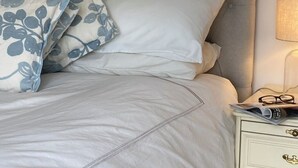 Ein ordentlich bezogenes Bett mit weißen und blauen blumigen Kissen, einem grauen Polsterkopfteil und einem Nachttisch mit Lampe und Zeitschrift.