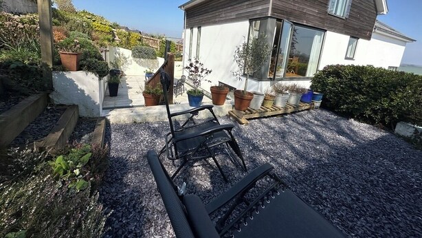 Ein modernes Haus mit Terrasse, ausgestattet mit einem schwarzen Liegestuhl und einem kleinen Tisch, umgeben von Topfpflanzen und einem Kiesweg.