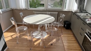 Et moderne køkken med et rundt bord og transparente stole, indbygget ovn og udsigt gennem store vinduer.