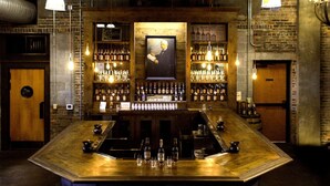 Un bar avec un comptoir en bois, des étagères remplies de bouteilles d’alcool, des ampoules suspendues et un tableau encadré au mur.