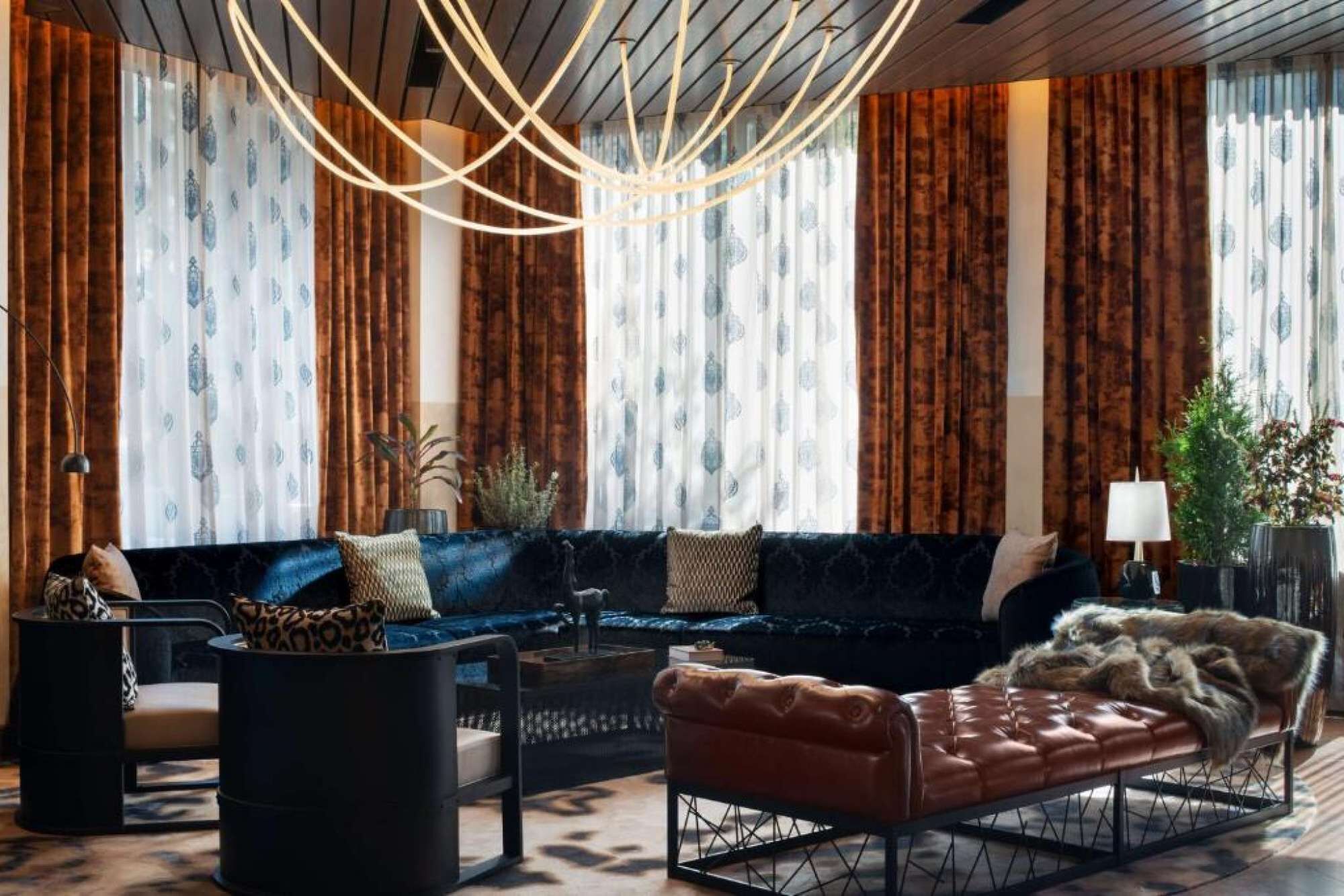 Ein modernes Wohnzimmer mit einem dunkelblauen Sofa, einer braunen Lederbank und einem Couchtisch. Vorhänge mit Blättermotiv, eine Stehlampe und ein Blumentopf sind ebenfalls vorhanden.