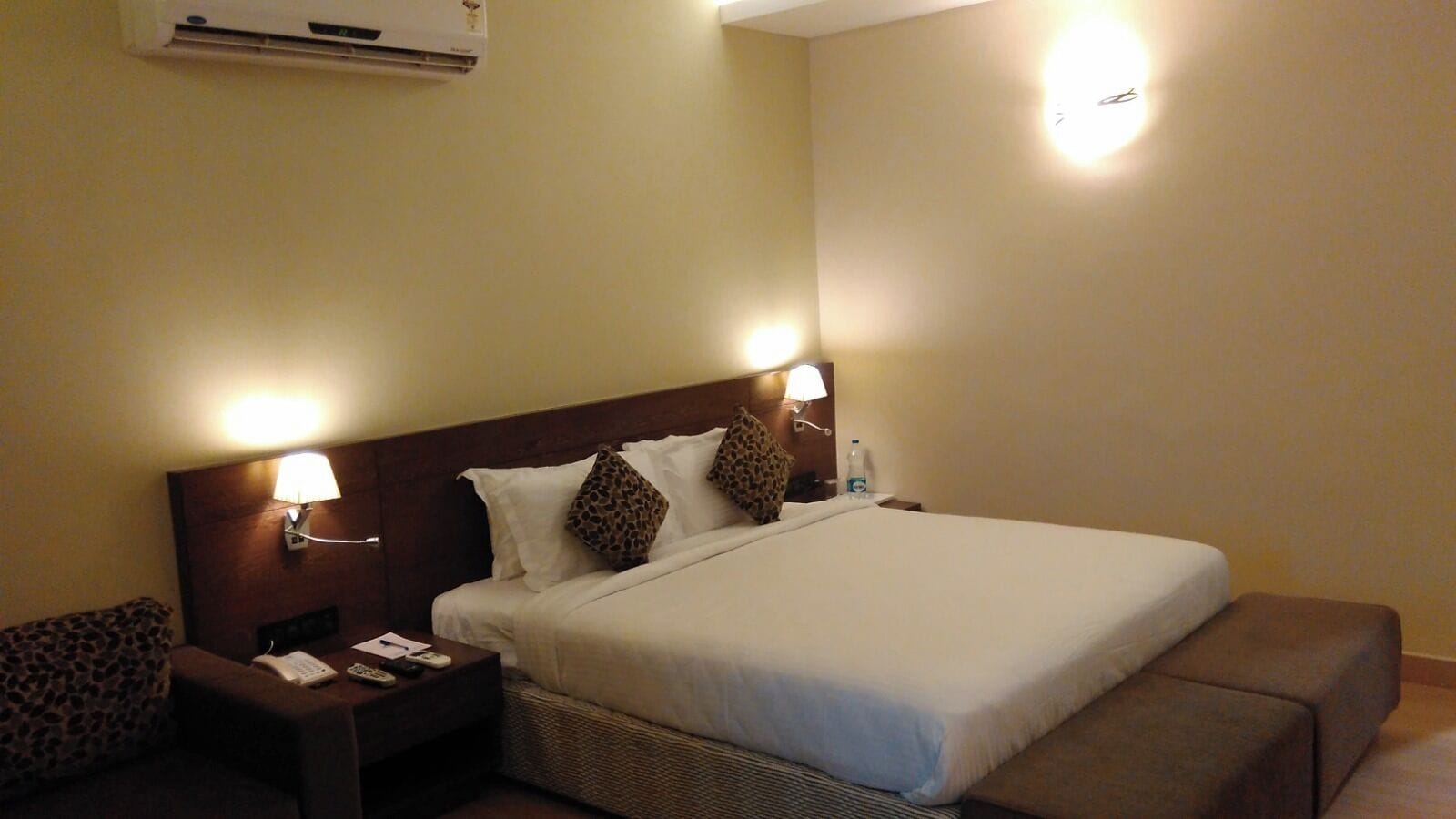 A hotel room with a large bed, two nightstands, a lamp, and a wall-mounted air conditioner.