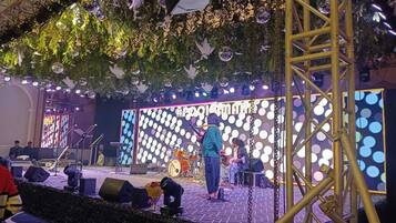 Un escenario con instrumentos musicales e intérpretes, rodeado de luces decorativas y follaje.