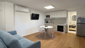 A modern living room with a grey sofa, a dining table with transparent chairs, a kitchen area with white cabinets, a flat-screen TV, and a built-in oven. - Villa Riviera 287 - Appartamento Pineta (Pescara)