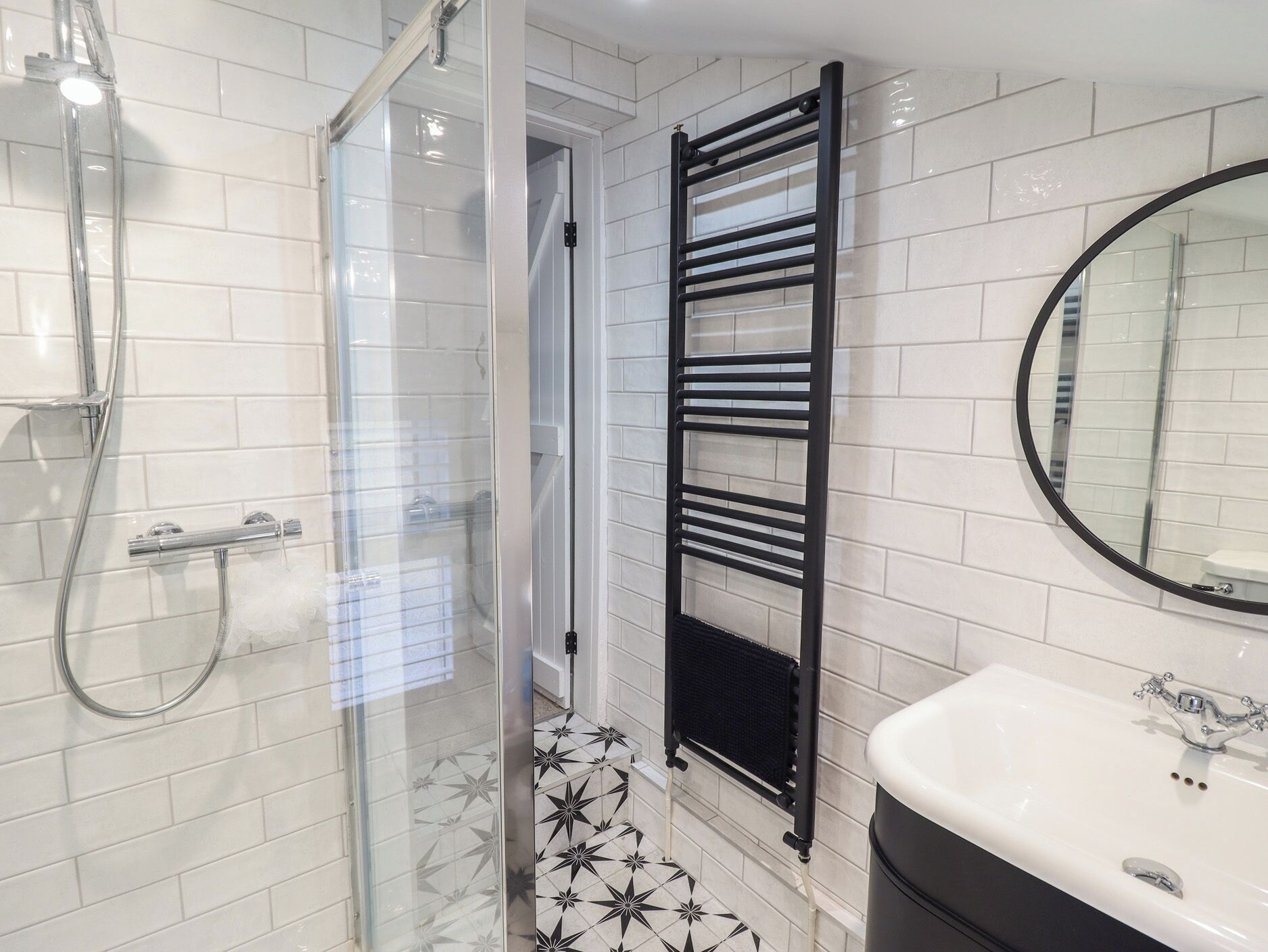 Un bagno con box doccia in vetro, porta asciugamani nero, lavandino bianco e specchio rotondo.