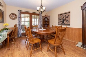 A dining room with a round wooden table, six wooden chairs, a chandelier, a wooden cabinet, and a wall-mounted clock. - Copper Lady Cottage - Creekside 3 BR, 2BA, Hot Tub, Firepit (Maggie Valley)