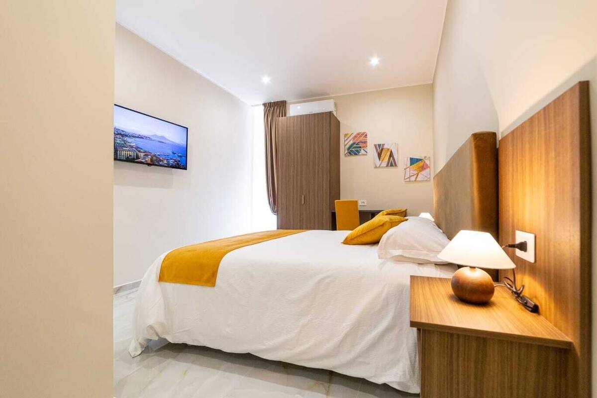 Habitació d'hotel amb un llit, una petita taula de nit amb una làmpada, un televisor a la pared i quadres murals.