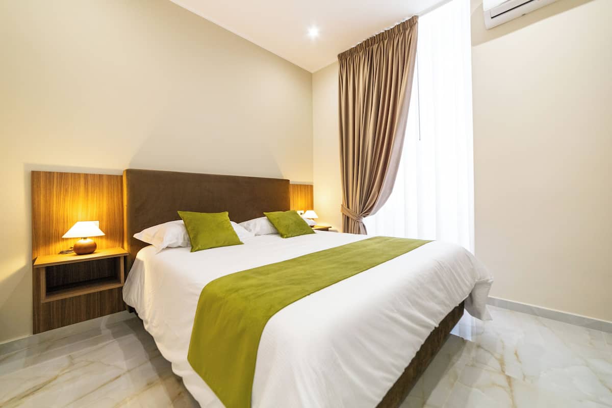 Habitació d'hotel amb un llit gran, una tauleta de nit amb una làmpada i una finestra amb cortines.