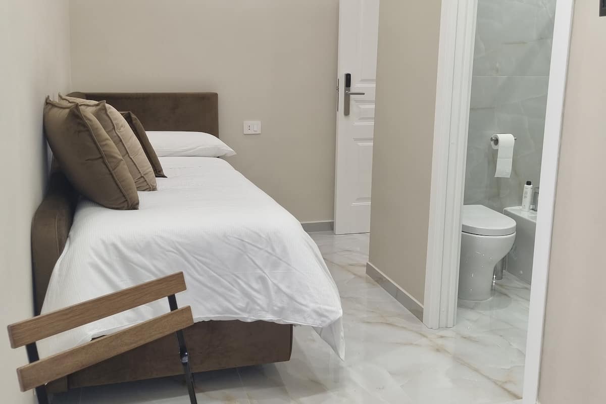 Habitació petita i neta, amb un llit, una banca de fusta i un bany visible a través d'una porta oberta.