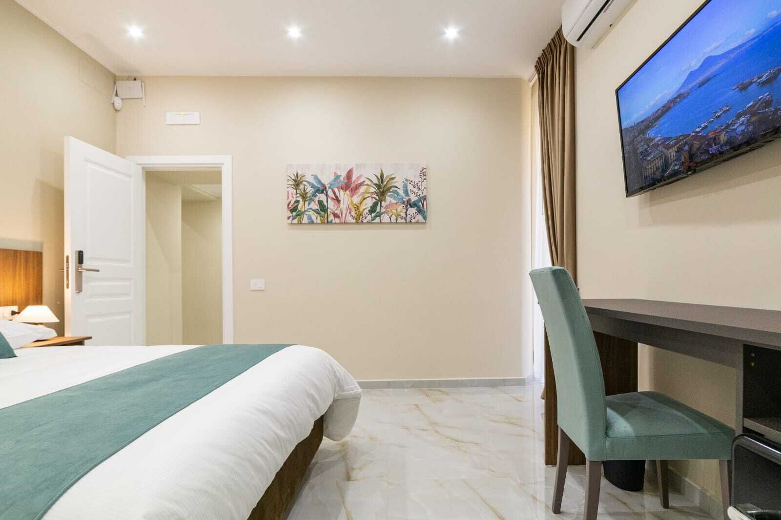 A hotel room with a bed, a desk, a chair, a painting on the wall, and a TV mounted on the wall.