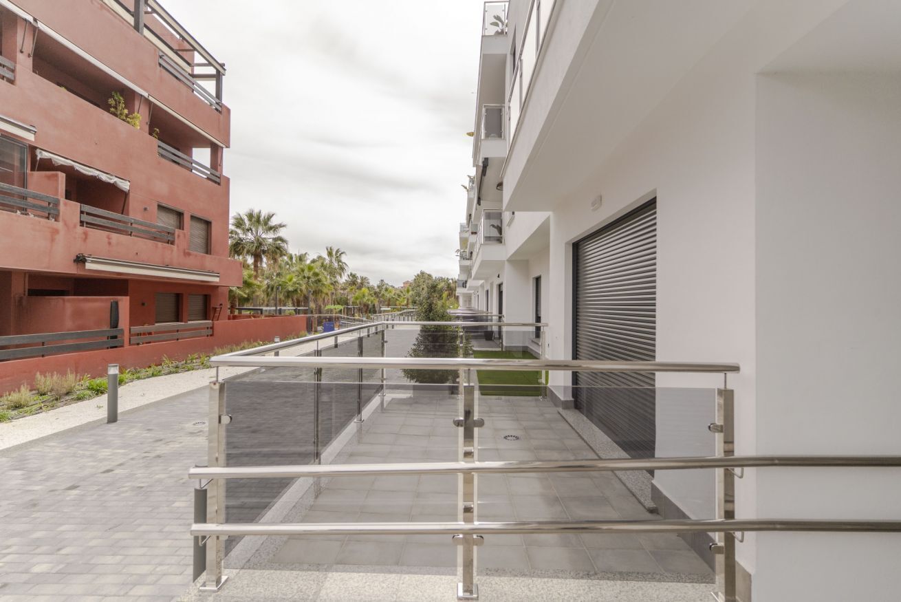 Un bâtiment moderne doté d’une passerelle avec des rampes en verre, d’un espace extérieur couvert et d’un palmier en arrière-plan.