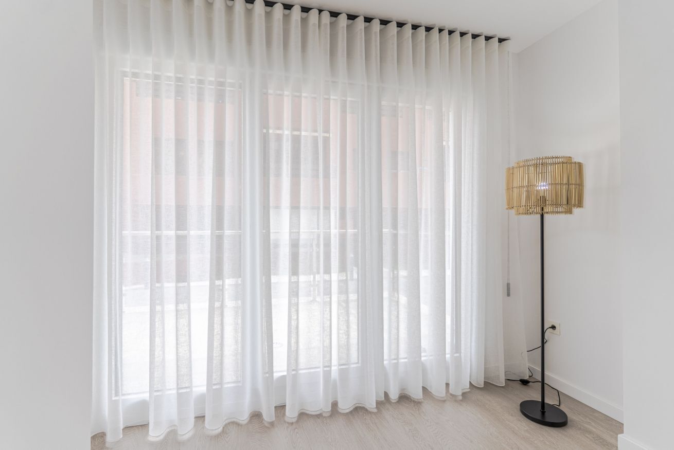 A room with sheer white curtains, a floor lamp, and a view of a building outside.