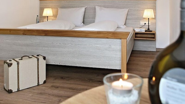 Quarto com estrutura de cama de madeira, duas mesas de cabeceira com abajures, uma mala e uma vela sobre uma mesa.