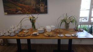 Uma mesa de jantar posta com pratos, tigelas e diversos alimentos, rodeada por vasos de flores e um quadro de uma igreja na parede.