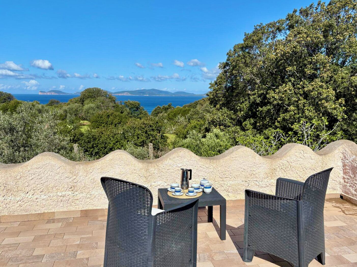 An outdoor seating area with wicker chairs and a table, overlooking a scenic view of the sea and hills.