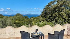 An outdoor seating area with wicker chairs and a table, overlooking a scenic view of the sea and hills.