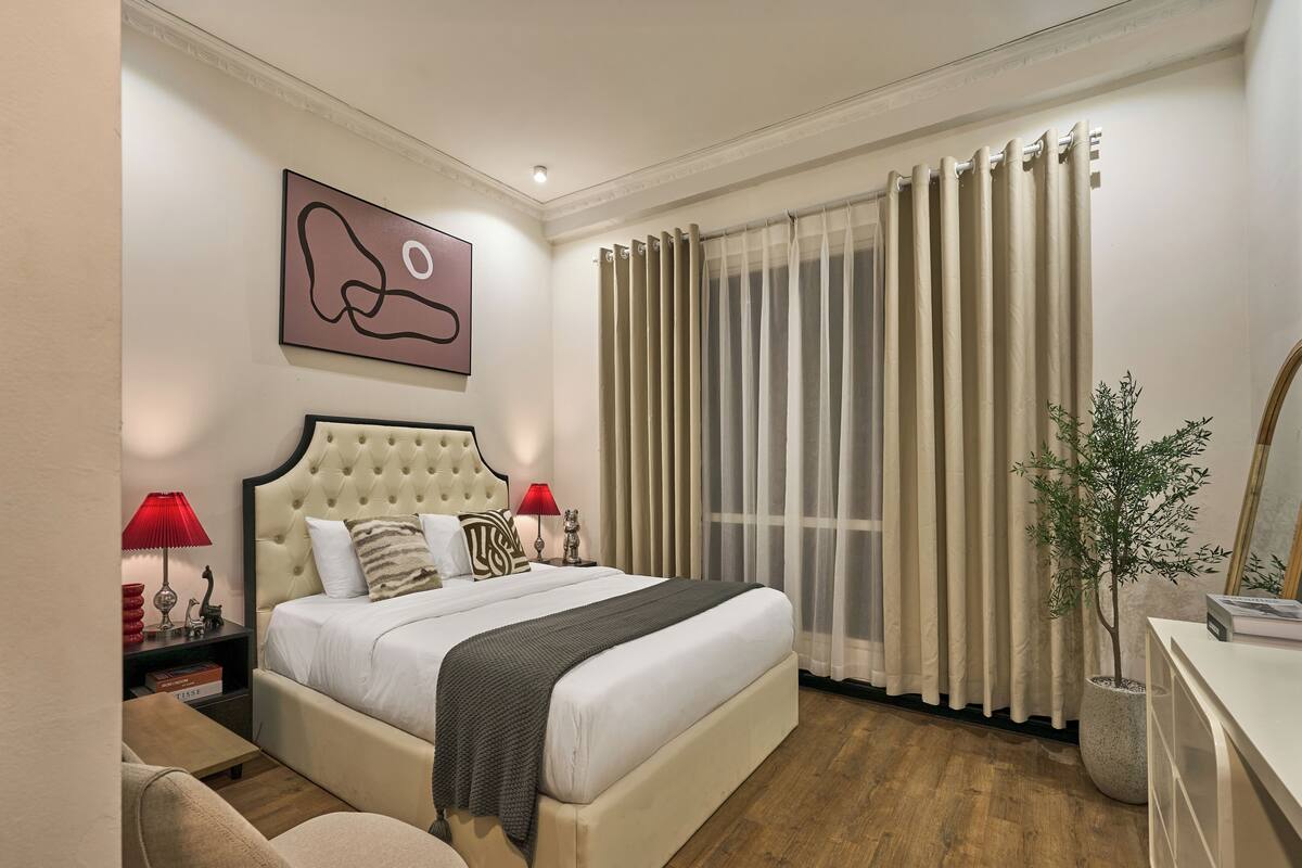 A hotel room with a large bed, two bedside tables with lamps, a wall-mounted artwork, a potted plant, and a large window with curtains.