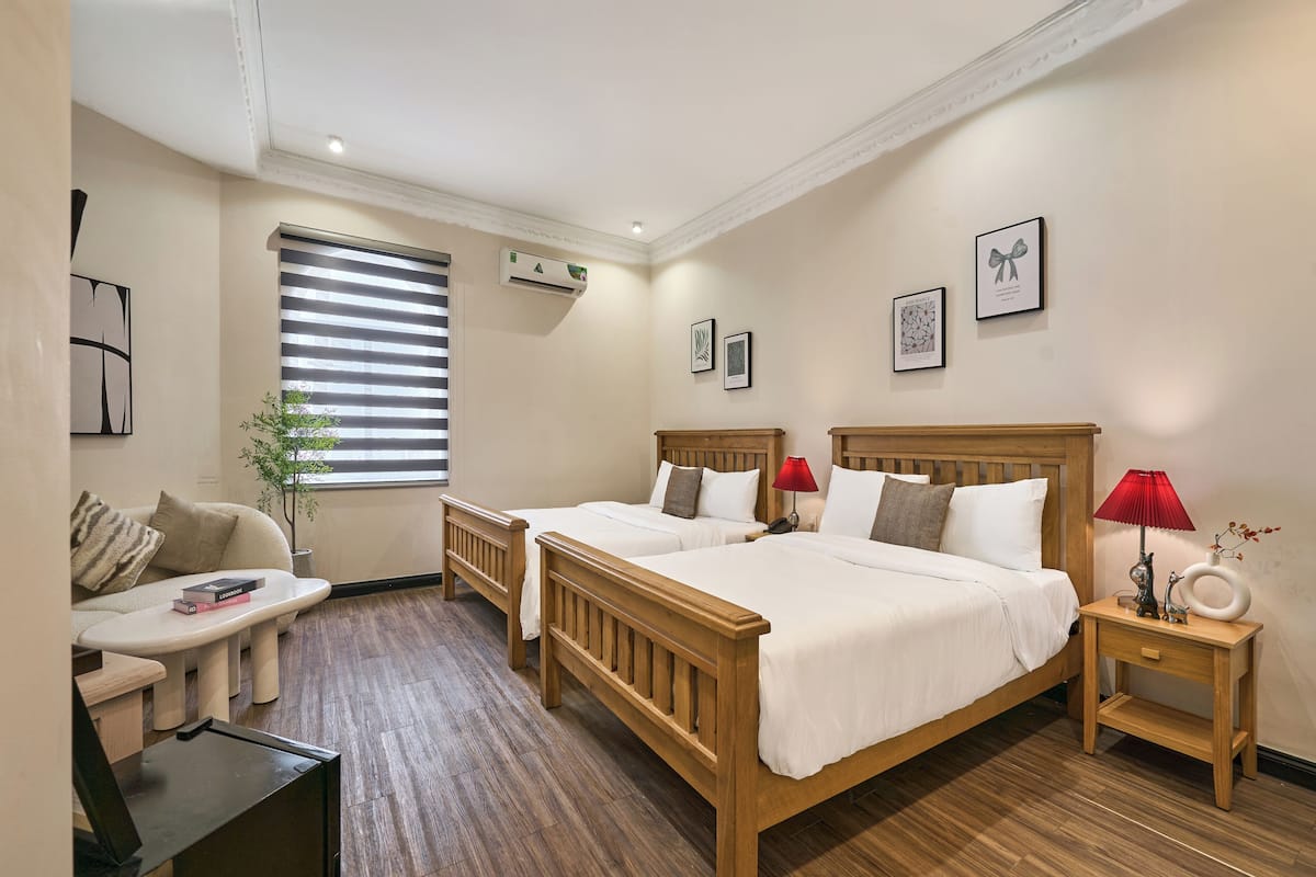 A hotel room with two beds, a sitting area with a table and chair, a window with blinds, and a small bedside table with a lamp and decorative items.