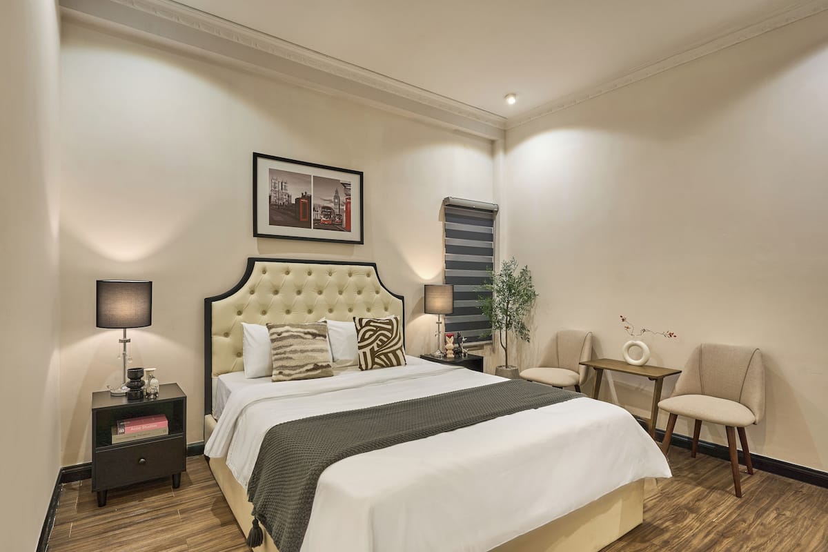 A hotel room with a large bed, two bedside tables with lamps, a small table with a chair, and a framed picture on the wall.