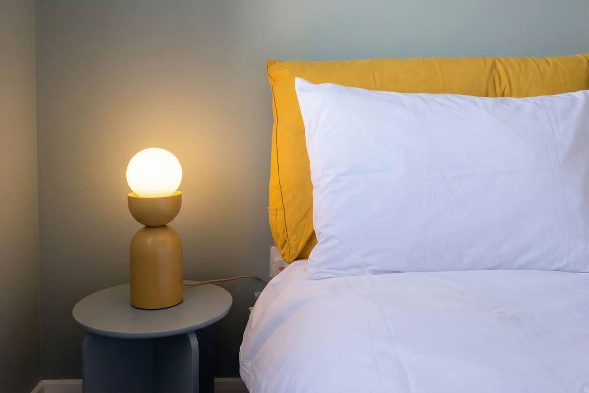 A bed with white bedding and a yellow pillow, a round table with a lamp, and a plain wall.