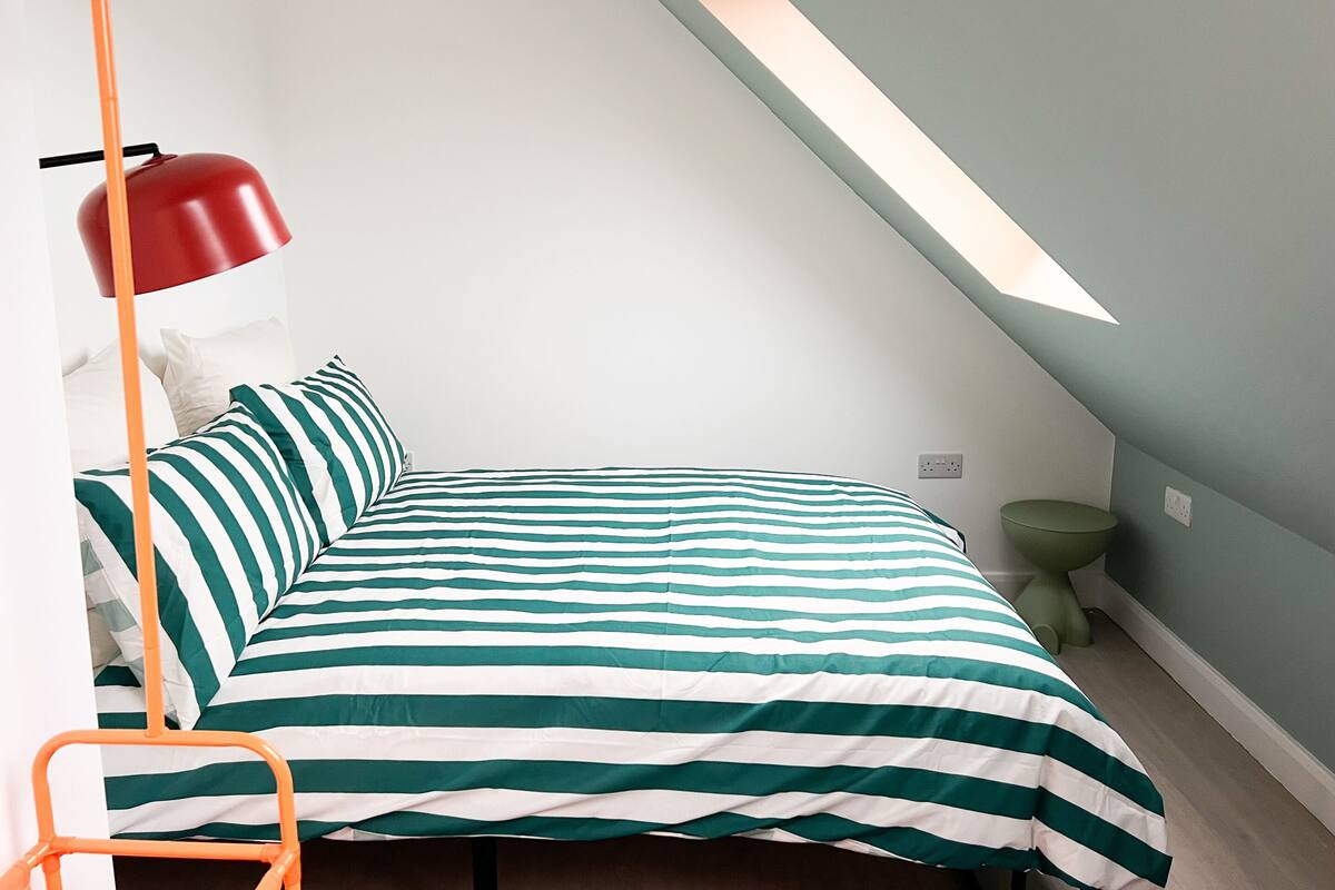 A bedroom with a bed, a red floor lamp, a green stool, and a skylight.