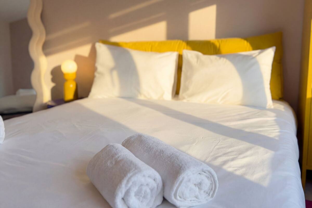 A neatly made bed with white linens and two rolled towels, a yellow headboard, and a bedside lamp.