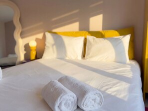 A neatly made bed with white linens and two rolled towels, a yellow headboard, and a bedside lamp. - Zozy Clitterhouse (London)