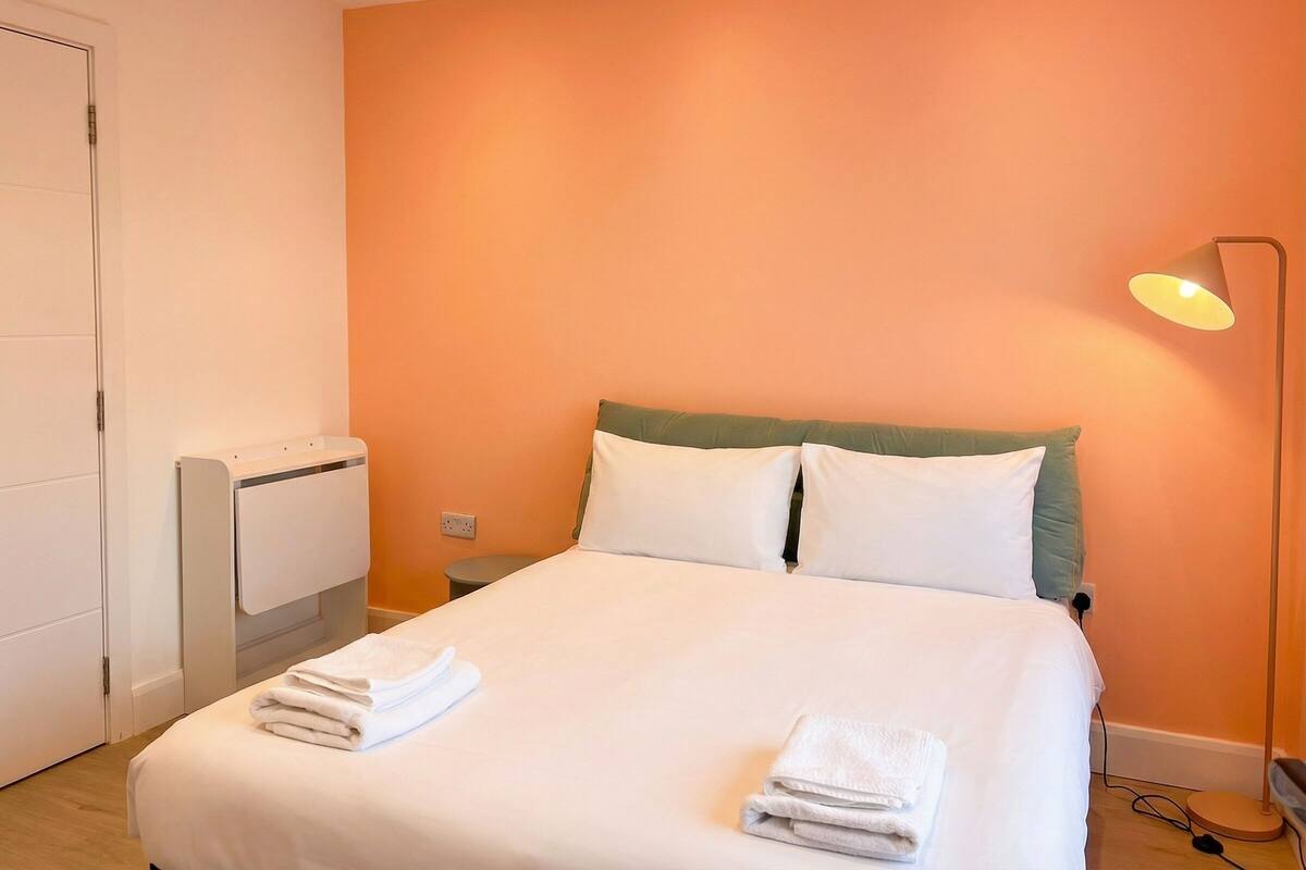 A neatly made bed with white linens and towels, a bedside lamp, and a radiator on the wall.
