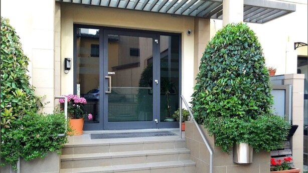 A modern building entrance with a glass door, steps leading up to it, and potted plants on either side. - 11. Appartmento (Signa)