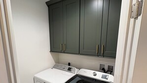 A compact laundry room with a front-loading washer and dryer, dark cabinets above, and a white door frame. - MSG host to confirm availability.• Spacious 3Bed 2BA •Sleeps 10 (Laie)