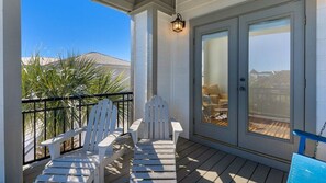 Un espacio exterior techado con dos sillas Adirondack blancas, una puerta de cristal que da a una habitación y una vista de un edificio al fondo.