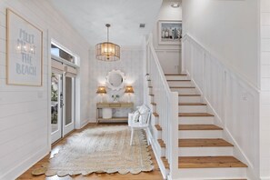 A hallway with a wooden floor, white walls, a staircase, a white chair, a mirror, and a rug. - 30A | 2 Mins to Beach | Private Pool & Beach! (Inlet Beach)