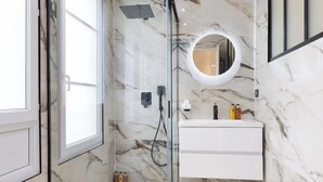 Un baño moderno con ducha de vidrio, tocador blanco con espejo circular y paredes de mármol.