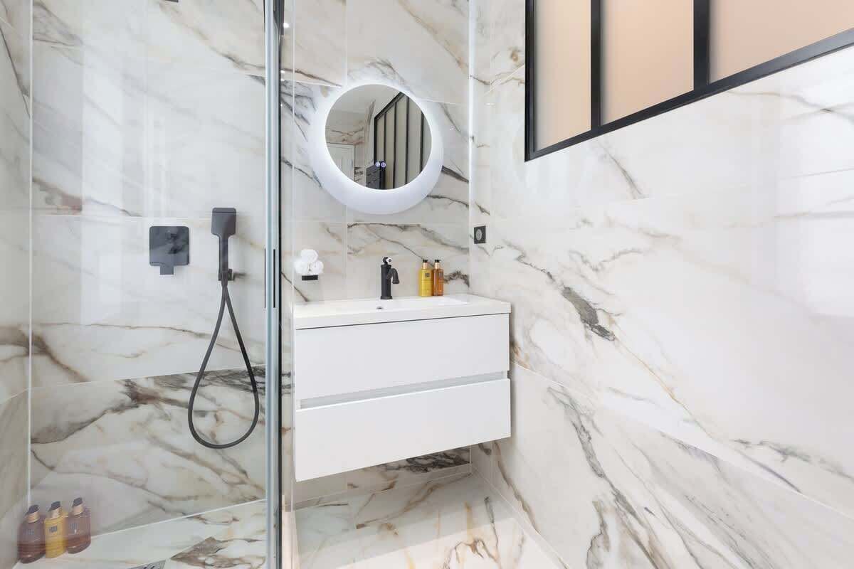 Ein modernes Badezimmer mit einer gläsernen Dusche, einem weißen Waschtisch mit Spiegel und Marmorwänden.