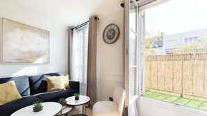 Una sala moderna con un sofá, dos mesas pequeñas, una silla, un reloj y un ventanal con vista a una cerca de bambú.