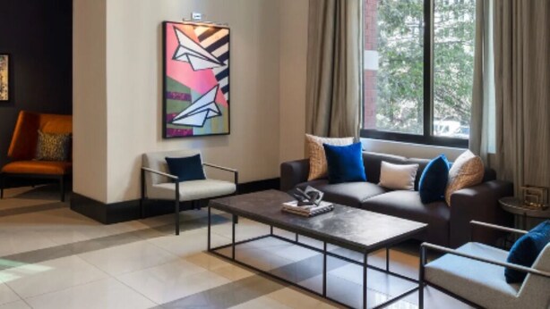 Uma sala de estar moderna com um sofá, poltronas e uma mesa de centro. Há um quadro na parede e uma janela com cortinas.
