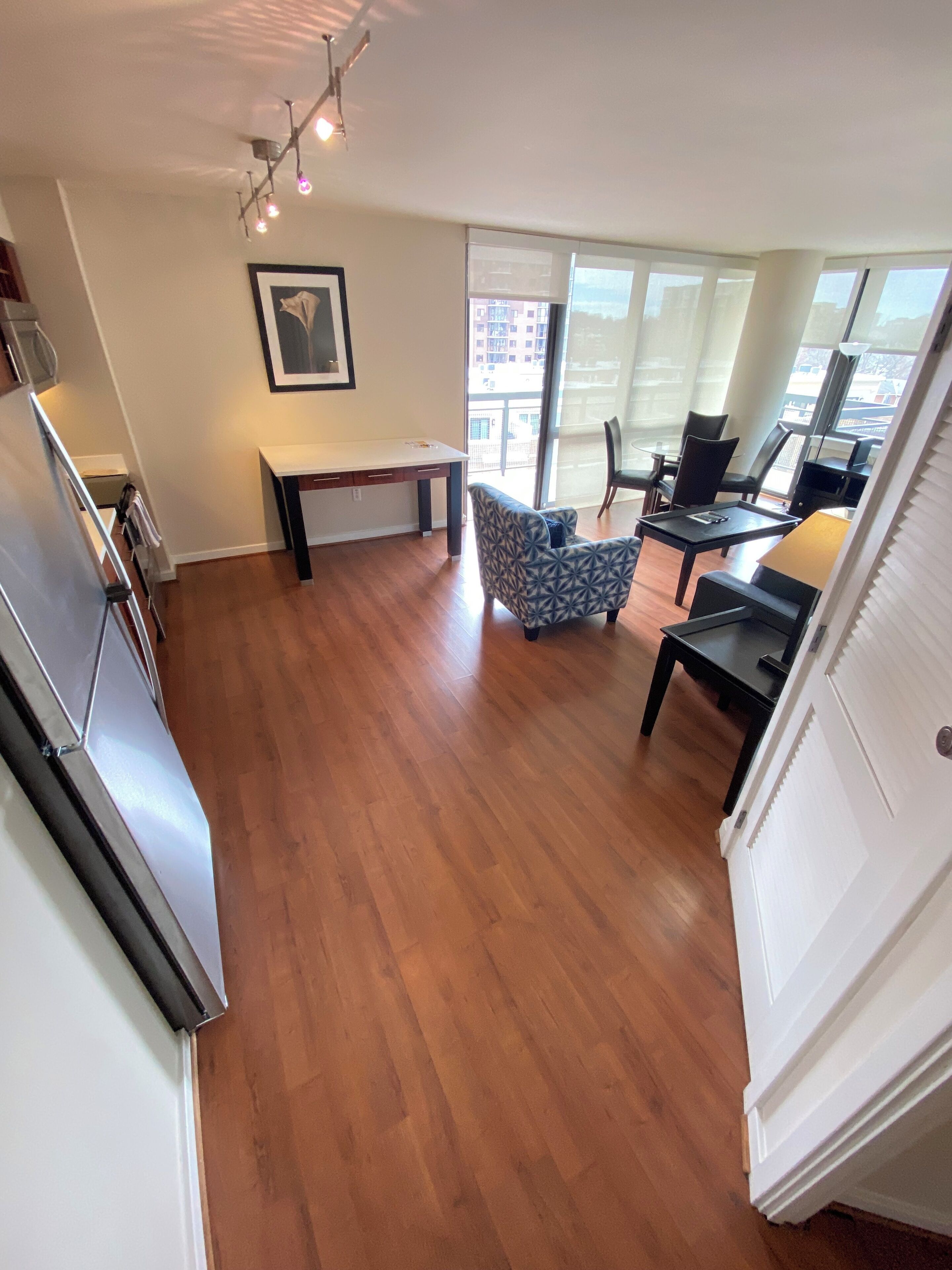 A spacious living area with a wooden floor, a dining table, and a sofa. There is a large window providing a view of the city.