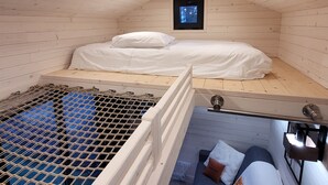 A small, cozy room with wooden walls and a sloped ceiling. It features a single bed with white bedding, a small window with a view of trees, and a metal grate on the floor. - The TINY HOUSE at Le Haras. (Avrillé)