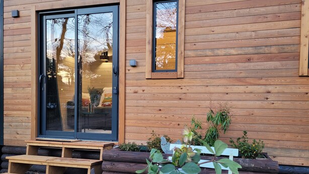 A wooden cabin with a sliding glass door, a small outdoor seating area, and a potted plant on a table. - The TINY HOUSE at Le Haras. (Avrillé)