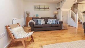Een ruime woonkamer met een houten vloer, een bruine leren bank, een houten stoel met een witte plaid en een trap naar een hoger niveau.