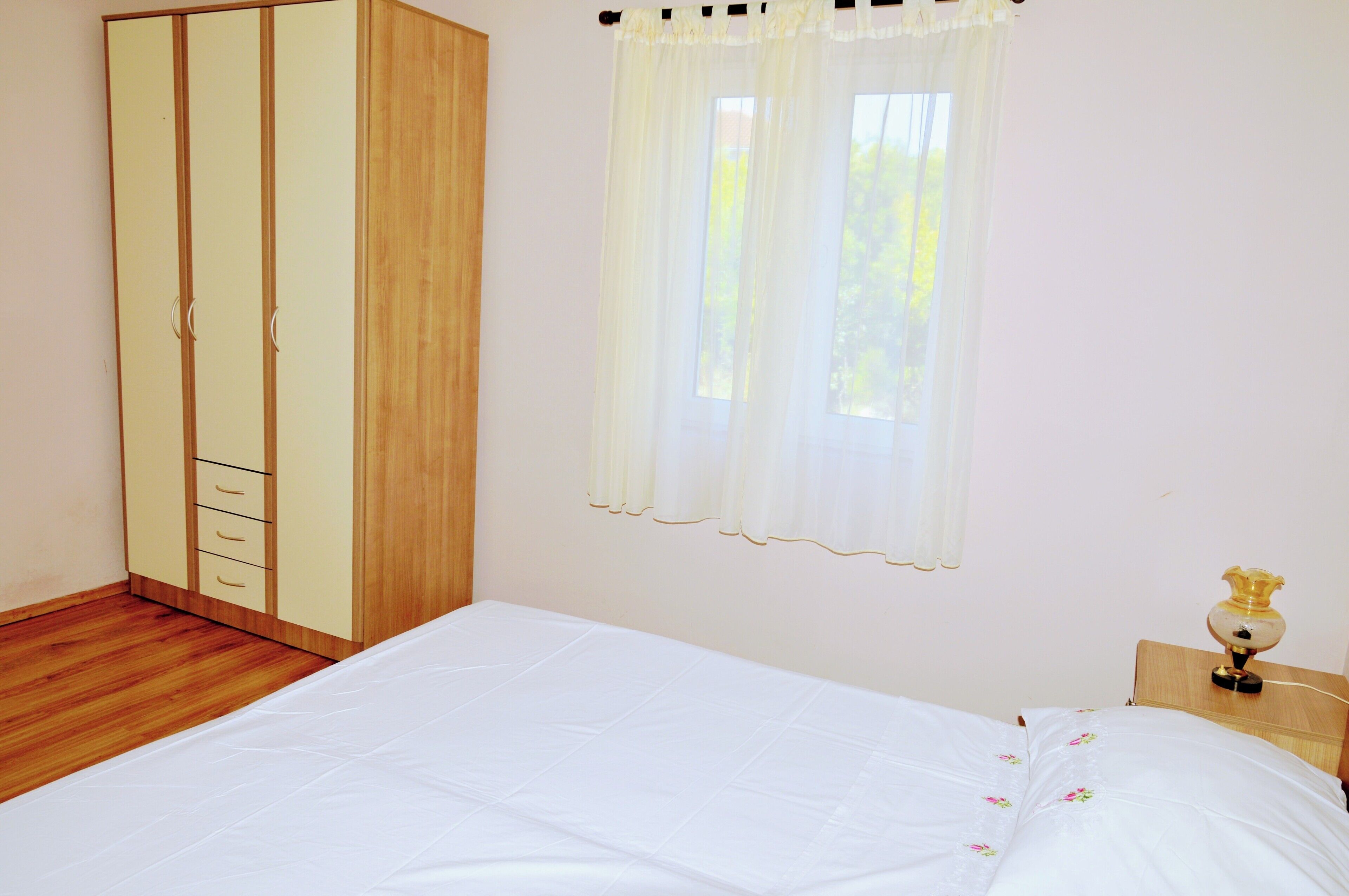 A bedroom with a wooden wardrobe, a bed with a white bedspread, and a window with sheer curtains.