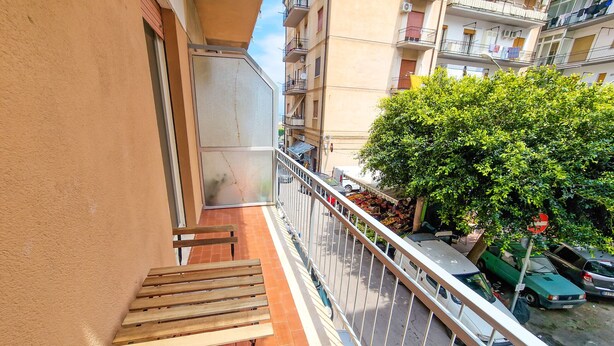 A balcony with a wooden bench and a white cushion, overlooking a street with parked cars and a tree. - Holy Dove - Giò Giò by Wonderful Italy (Cefalù)