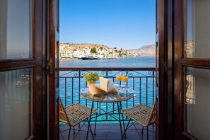 Un balcone con sedie in vimini, un tavolino di vetro con un libro e due bicchieri di succo d'arancia, e vista su una località costiera attraverso l'acqua.
