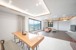 A modern living room with a wooden dining table, a sofa, a TV, and a large window offering a city view. - hotel onggi (Gyeongju)