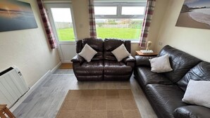 A living room with two leather sofas, a brown rug, and a view of a green landscape through a window. - Atlantic View Chalets #2 | Self-catering holiday cottage near Crantock beach (Crantock)