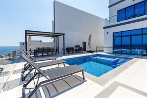 A rooftop pool area with sun loungers, a barbecue, and a glass balustrade overlooking the sea.