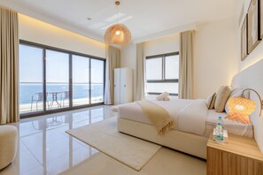 A modern hotel room with a large bed, a wooden nightstand, a glass-enclosed balcony with a view of the sea, and a large window with curtains.