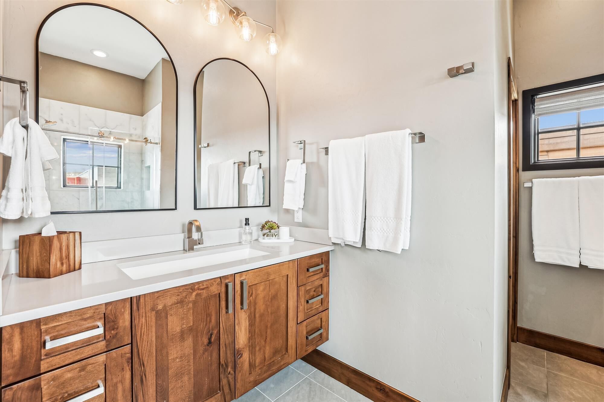 Un bagno moderno con un mobile bagno in legno, due ampi specchi, una finestra e un porta asciugamani.