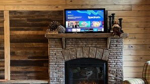 A cozy living room with a stone fireplace, a television mounted above it, and a bowl of pinecones on the floor.