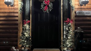 A log cabin entrance with a black door, two Christmas trees, a wreath, and a welcome mat.